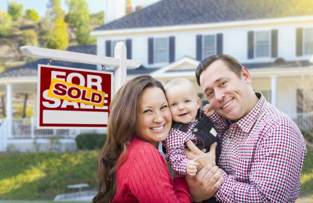 Happy Young Family In Front Of For Sale Real Estate Sign And House.